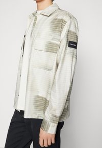 Camisa abotonada en tono beige claro con patrón de cuadrícula, dos bolsillos frontales y marca Calvin Klein en la manga. Textura de tela suave.