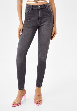Femme portant un jean skinny noir délavé taille haute et des sandales roses à talons hauts et brides, debout devant un fond blanc.