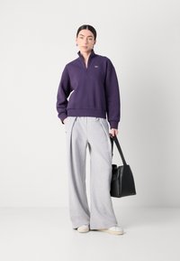 Paarse kwart-rits sweatshirt met geribbelde zoom, gecombineerd met lichtgrijze wijde sweatpants en een zwarte totebag; model draagt witte schoenen.