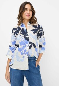 Button-up shirt in light fabric with large blue and black floral pattern. Features a collared neck and rolled-up sleeves. Paired with blue pants.