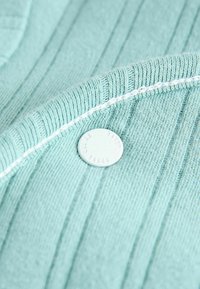 Jasnoniebieska prążkowana dzianina o gładkiej fakturze, z białym guzikiem z wytłoczonym napisem. Akcent białego szwu wzdłuż krawędzi.