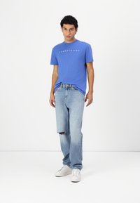 Lys blå t-shirt med "TOMMY JEANS" logo, kombineret med slidte blå jeans med et hul ved knæet. Hvite sneakers fuldender outfittet.