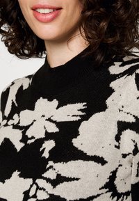 Tröja med ett blommönster i svart och krämfärgat, med en texturerad stickad kvalitet och en hög, ribbad krage. Närbild visar designens detaljer.