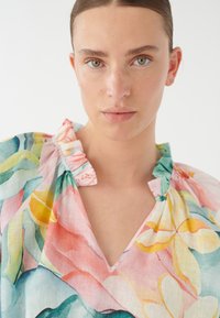 Blusa in tessuto leggero con fantasia floreale colorata, caratterizzata da scollatura a V e colletto a balze. I colori predominanti sono il rosa, il verde e il giallo.