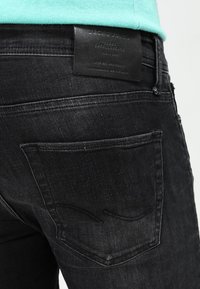 Mustad denim püksid, millel on tekstureeritud pind, nähtavad tagapocketi õmblused ja nahk silt, millel on kirjas "Jack & Jones" vöökoha juures.