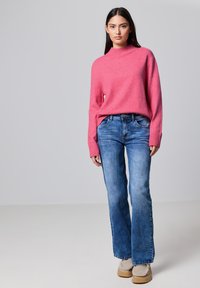Maglione rosa a maglia con collo alto, abbinato a jeans svasati chiari. La modella indossa scarpe beige con una finitura testurizzata. Look semplice e moderno.