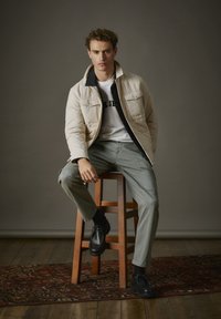Padded cream jacket with black collar, worn over white t-shirt, paired with olive green cargo pants and black shoes, seated on a wooden stool.