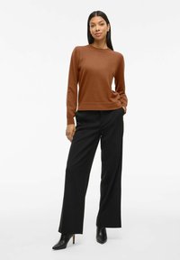 Pull en tricot marron avec des manches longues, un col rond, associé à un pantalon large noir et des bottes à talons noires. Texture lisse.