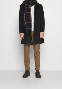 Zwarte jas, gebreide crèmekleurige trui, bruine broek, bruine leren laarzen en een marineblauw geruite sjaal met franjes, gedragen door een staande persoon.