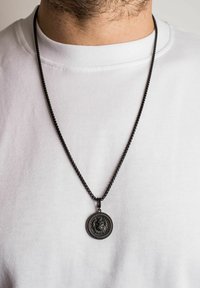 Sort metalkæde med en cirkulær vedhæng, der har et intrikat løvehoveddesign og tekstureret kant, båret over en hvid skjorte.