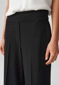 Black, high-waisted trousers made from a smooth, structured fabric with a front pleat detail and a wide, straight-leg design.