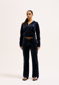 Marinevelours trainingspak met een zip-up hoodie en broek met een trekkoord. Glad textuur met subtiele branding op de hoodie. Gecombineerd met slippers.