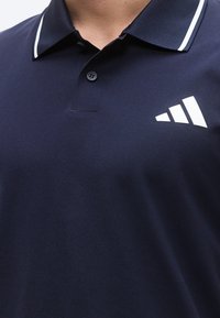 Μπλε ναυτικό πόλο πουκάμισο με γιακά, λευκές διακοσμητικές ρίγες και λευκό τρίγωνο λογότυπο στο αριστερό στήθος. Υφή υφάσματος.