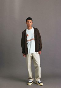 Brown knitted cardigan over a white graphic tee, beige trousers, and black and yellow sneakers. Standing with a neutral backdrop.