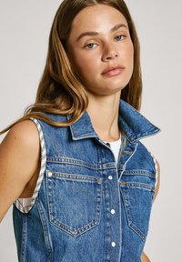 Džínová bezrukávová bunda svetlomodrej farby s kontrastnou pruhovanou látkou na pleciach, má predné vrecká a zapínanie na cvoky.