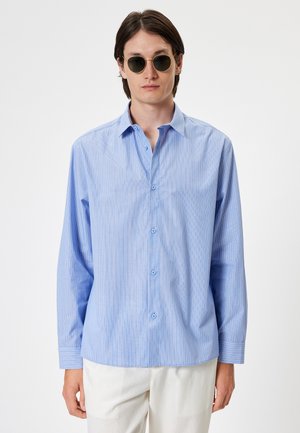 Camisa a rayas azul claro con botón frontal y mangas largas. La tela parece suave y los botones son de un azul a juego.