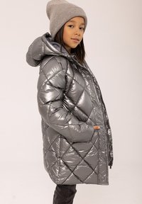 Sudraba pūkaina jaka ar dimanta quiltu rakstu, kapuci un rāvējslēdzēju. Siltīgs, spīdīgs materiāls, valkāts virs tumšiem biksēm un pelēka adīta cepure.