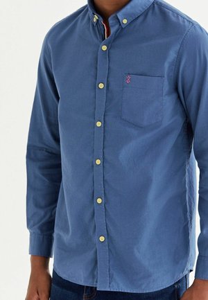Hombre con camisa azul de manga larga abotonada, con bolsillo en el pecho y botones amarillos, mostrado desde el cuello hasta las caderas.