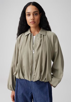 Frau mit langen, lockigen Haaren, die eine hellolivfarbene, mit Reißverschluss versehene Jacke mit Kragen und weiten Ärmeln trägt, kombiniert mit blauen Hosen.