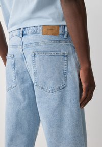 Jean en denim bleu clair avec une coupe décontractée, doté de deux poches arrière et d'une étiquette en cuir tan à la taille portant l'inscription "DENIM SUPPLY".