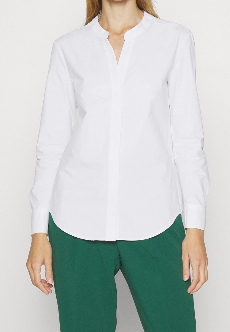 Femme portant un chemisier blanc à manches longues avec col mandarin et un pantalon vert sur un fond uni clair.