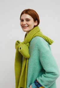 Grüner gestrickter Schal mit einer Schleifendetail, getragen über einem hellblauen flauschigen Pullover. Der Schal zeichnet sich durch ein strukturiertes, geripptes Muster aus.