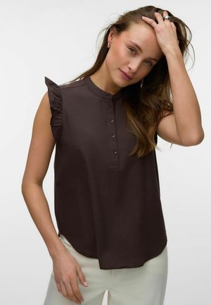 Kobieta z długimi brązowymi włosami dotykająca głowy, ubrana w ciemnobrązową bluzkę bez rękawów z falbankami na ramionach i jasne spodnie.