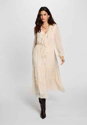 Donna con capelli lunghi e scuri che indossa un vestito midi beige con volant e stivali neri alti fino al ginocchio, in piedi contro uno sfondo chiaro e uniforme.