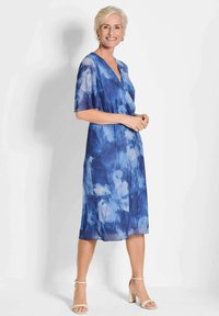 Blaues Blumenkleid aus leichtem Stoff, mit V-Ausschnitt, kurzen Ärmeln und fließender Silhouette. Akzentuiert mit hellen Schuhen.