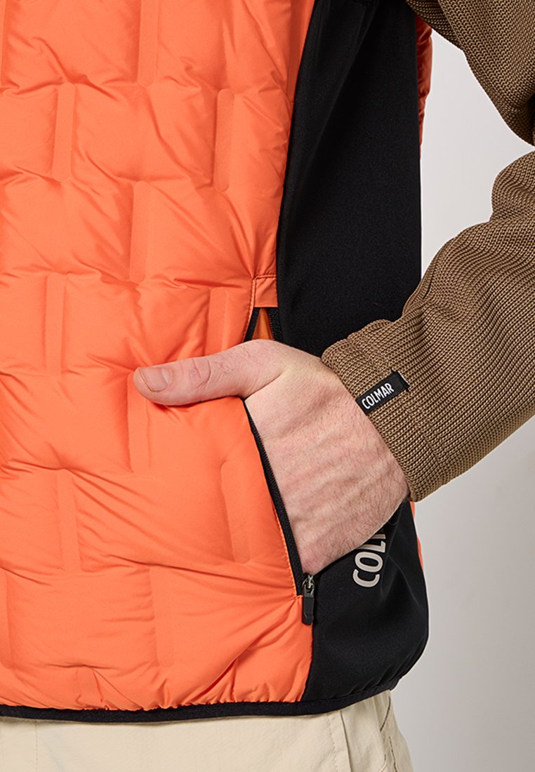 Gepolsterte orange Weste mit einem schwarzen unteren Abschnitt. Verfügt über eine Reißverschlusstasche, einen strukturierten braunen Ärmel und ein Logo an der Seite.