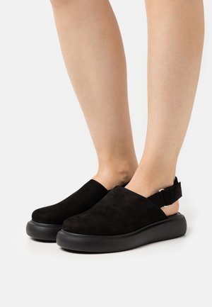 Chaussures slingback en suède noir avec un bout arrondi, une semelle plate en caoutchouc et une bride réglable au talon, présentant une texture lisse.