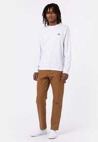 Witte lange mouwen shirt met een klein gekleurd logo op de borst, gecombineerd met bruin rechte broek en witte sneakers.