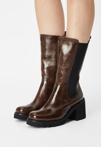 Bottes chevilles en cuir marron avec une finition brillante, des panneaux élastiques sur les côtés et un talon bloc. Elles comportent une fermeture éclair et une semelle texturée.