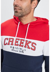 Felpa con cappuccio dal design colorblock rosso, bianco e blu navy. Presenta una tasca frontale, lacci e il testo "CREEKS ORIGINAL Dal 1974".