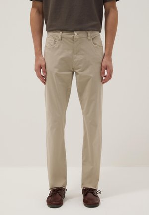 Uomo con pantaloni beige a gamba dritta, scarpe marrone scuro e camicia scura a maniche corte, in piedi davanti a uno sfondo semplice.
