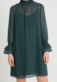 Tikkusävyinen, läpikuultava georgette-mekko, jossa on laikullinen kuvio, röyhelökaulus ja levenevät hihat. Sujuva rakenne, löysä malli, ulottuu reiden keskiosaan.