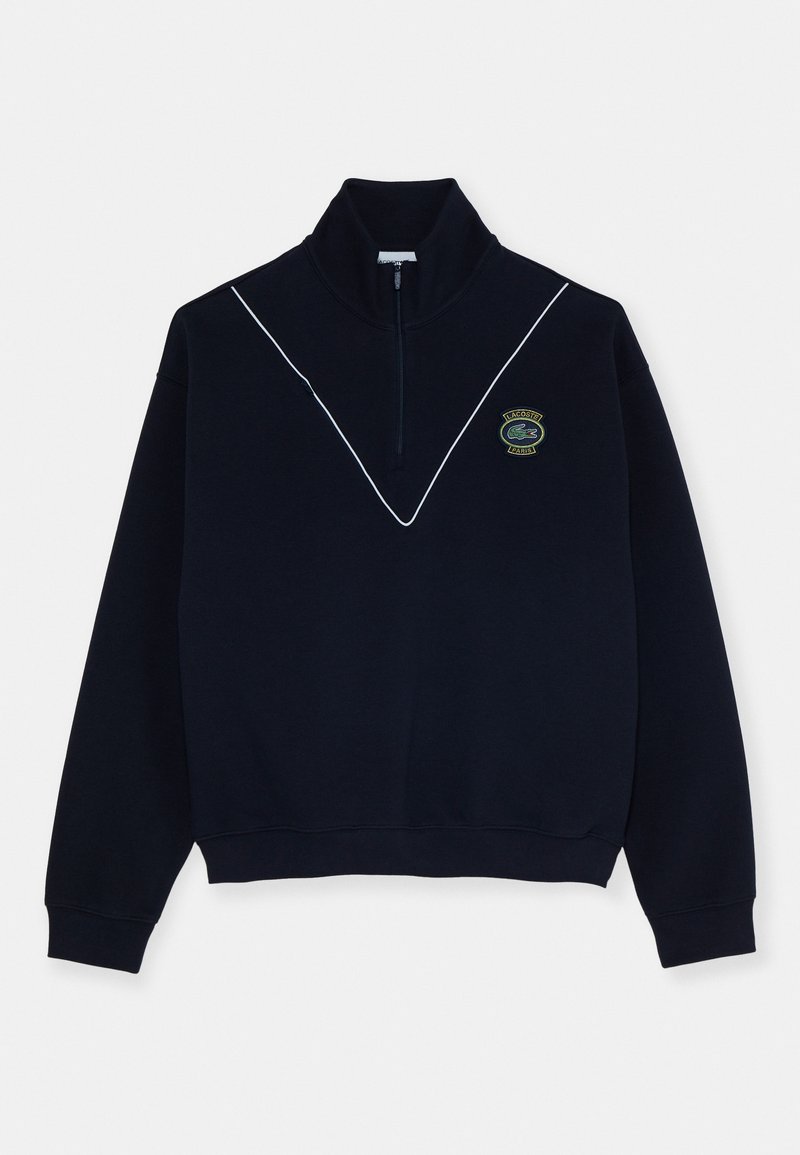 Sweatshirt bleu marine zippé avec des garnitures blanches formant une forme en V et un patch logo crocodile de Lacoste Paris sur la poitrine gauche.