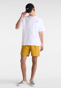 Weißes Kurzarm-T-Shirt mit rotem und blauem "VANS OFF THE WALL"-Text, kombiniert mit gelben Shorts und schwarz-weißen karierten Slip-On-Schuhen.