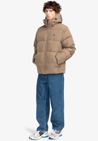 Giacca imbottita beige con colletto alto, chiusura con zip, tasche laterali e polsini elastici, abbinata a jeans blu e sneakers bianco e nere.