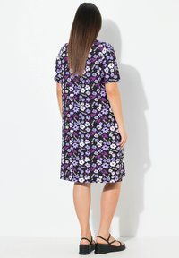 Mustat lyhythihaiset mekko, jossa on valkoisia ja purppuranvärisiä kukkakuoseja, löysä malli ja polvipituinen muotoilu. Korostettu litteillä sandaaleilla.