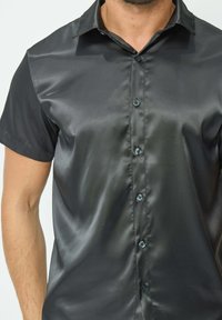 Chemise noire à manches courtes avec des boutons, confectionnée dans un tissu lisse et brillant. Présente un col classique et six boutons assortis sur le devant.