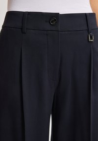 Zwarte op maat gemaakte broek met plooien, riemlussen, knoopsluiting en een klein metalen detail aan de rechterzijde bij de tailleband.