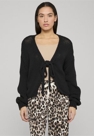 Cardigan noir en maille avec fermeture à nouer sur le devant, décolleté en V profond et manches longues et volumineuses. Porté sur un haut beige et un pantalon à imprimé animal.
