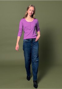 Lila pitkähihaisen paita, jossa on nappisuljenta kaula-aukoissa, yhdistettynä sinisiin farkkuihin ja mustiin nilkkakorkokenkiin, vaaleanvihreää taustaa vasten.