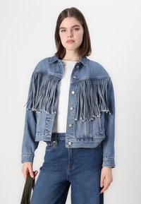 Blaue Jeansjacke mit langen Fransen, Knopfverschluss und zwei Vorderaschen, kombiniert mit einer engen blauen Jeans und einem weißen Oberteil.