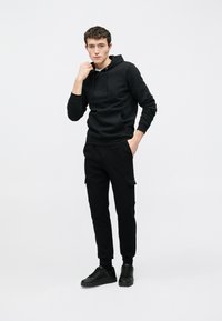 Fekete kapucnis pulóver elülső zsebbel és állítható húzózsinórral, fekete cargo nadrággal, oldalzsebekkel, valamint fekete sportcipővel párosítva.