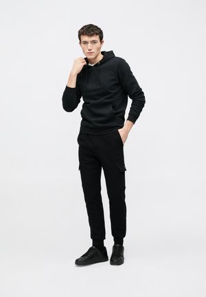 Musta huppari, jossa on etutasku ja säädettävä kiristysnyöri, yhdistettynä mustiin cargo-housuihin sivutaskuilla ja mustiin lenkkareihin.