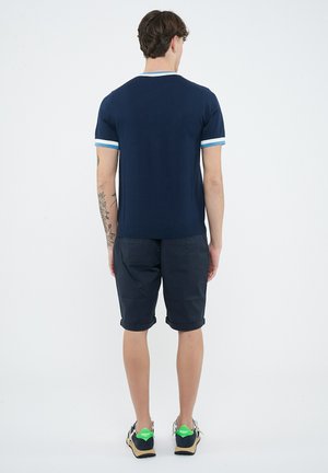 Uomo in piedi di spalle che indossa una maglietta blu navy con bordi azzurri, pantaloncini scuri, scarpe da ginnastica e con un tatuaggio visibile sul braccio, su sfondo bianco.