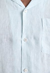 Gaiši zils lina krekls, īss apkakle, priekšpuses pogas, vienāda krūšu kabata, teksturēta auduma ar smalku austumu rakstu.