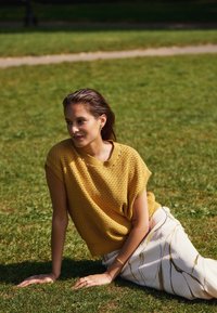 Žlutý pletený svetr s krátkým rukávem, zkombinovaný s bílou sukní s zlatými a černými pruhy. Pozadí tvoří tráva, uvolněná póza.